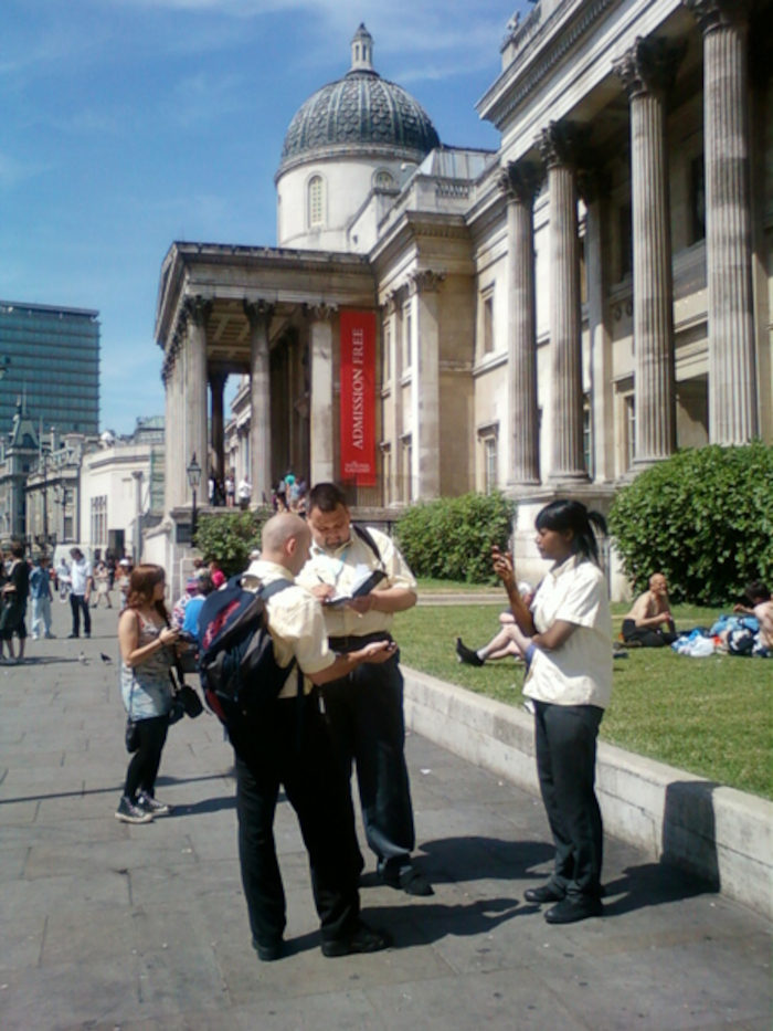 Group of people, predominantly men, standing in front of a classical building, possibly a museum or gallery. One man appears to be consulting a document with another. A woman in a white shirt is standing slightly apart, seemingly engaging with her phone. Other individuals are visible in the background, some seated on the grass. A banner reading Admission Free is displayed on the building. The scene is set outdoors on a sunny day. The overall impression is of a casual gathering possibly near a cultural institution.