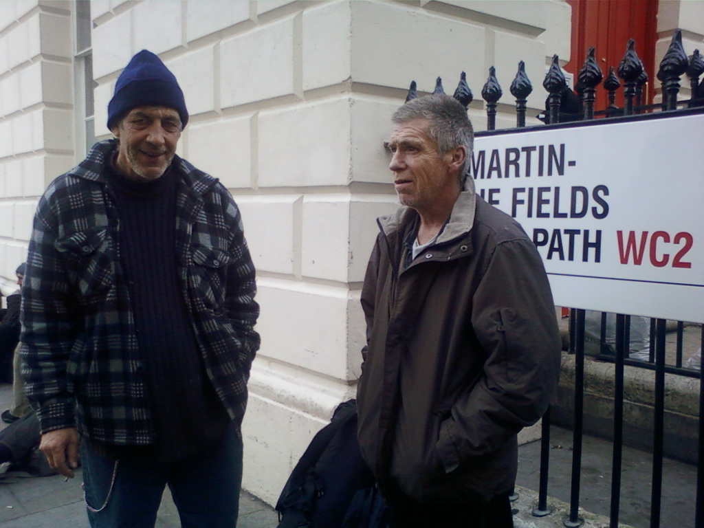 Two men standing next to a street sign that reads Martin-e Fields Path WC2. Both men appear to be of middle age or older and are dressed in casual, somewhat worn clothing. The man on the left wears a plaid shirt and a beanie, while the man on the right wears a dark-colored jacket. The background is a white building with a section of red visible on the right. The overall impression is one of an informal, possibly candid street scene. The setting suggests a possibly urban environment.