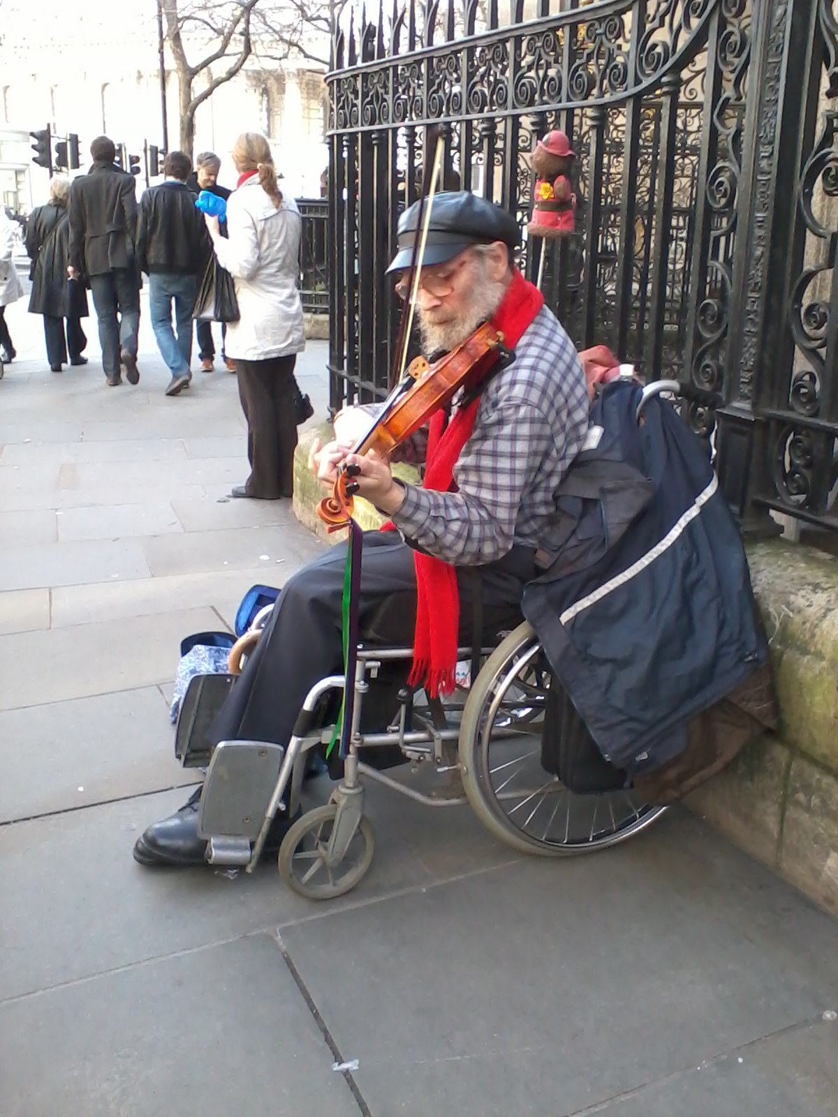 Man in a wheelchair playing the violin on a city street. He is wearing a plaid shirt, a red scarf, and a cap, and appears to be busking. Behind him is an ornate metal fence and other pedestrians are visible in the background, suggesting a public urban setting. The overall mood is one of quiet dignity and perseverance.