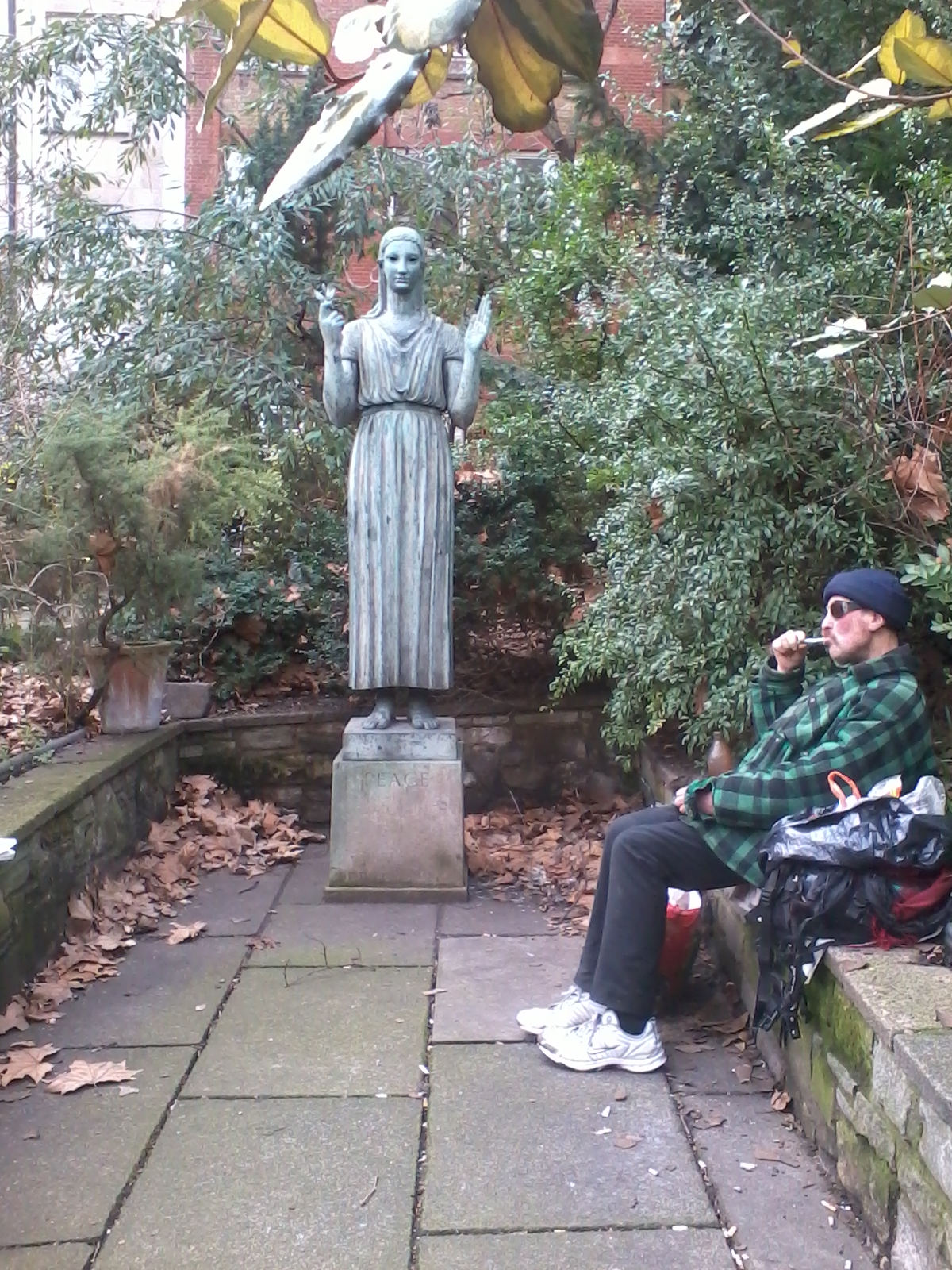 Statue of a woman, possibly representing Peace, as indicated by the inscription on the pedestal. The statue is situated in a small, enclosed garden setting with stone paving and overgrown greenery. A man, dressed in casual clothing and wearing sunglasses, sits on a low wall in the garden, seemingly observing or contemplating the statue. The overall mood is quiet and contemplative. The juxtaposition of the classical statue and the contemporary man suggests themes of time, reflection, and contrast between the ideals represented by the statue and the reality of everyday life.