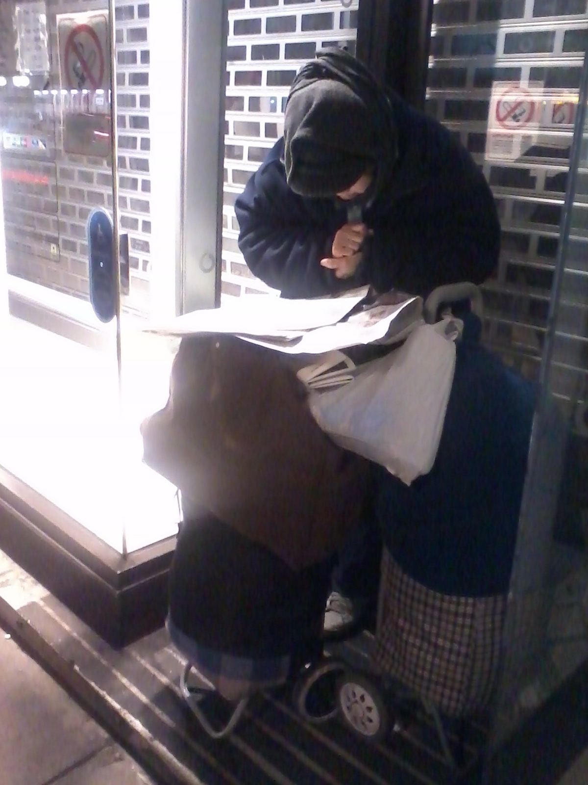 Person, heavily bundled in dark clothing and a hooded sweatshirt, hunched over what appears to be newspapers or papers resting on a small wheeled cart. They are seated in a doorway, possibly seeking shelter. The overall impression is one of poverty or homelessness. The scene is dimly lit, adding to the sense of bleakness.