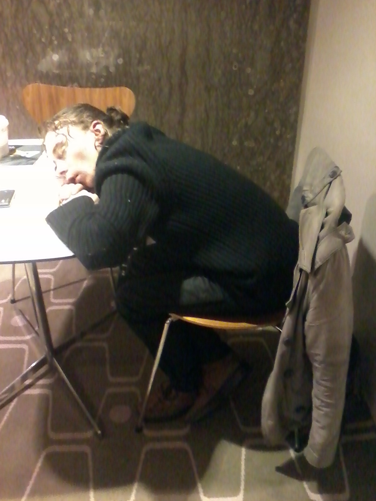 Person sleeping with their head resting on a white table, their body slumped over a chair. The person is wearing a dark sweater and pants, and their hair is pulled back. A light-colored jacket is draped over the back of the chair. The background is a mottled brown wall, and the floor is covered in a patterned carpet. The overall impression is one of weariness or exhaustion.