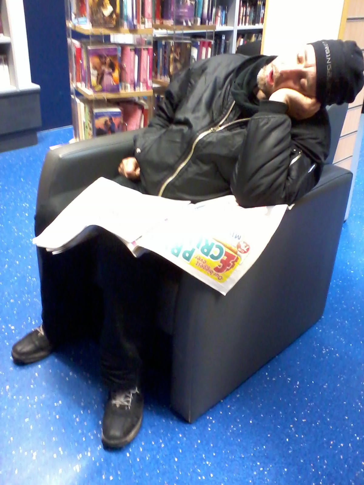 Man sleeping in an armchair in what appears to be a library.  He's wearing a black jacket and hat, and has a newspaper or magazine resting on his lap. The library shelves are visible in the background. The overall impression is one of quiet exhaustion or weariness.
