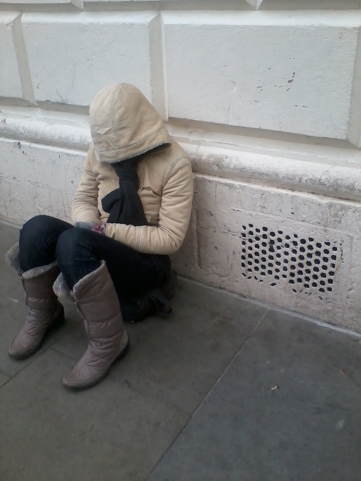 Person sitting on the ground, huddled in a beige hooded jacket and dark-coloured pants and boots. Their head is lowered, concealed by the hood, suggesting a posture of dejection, vulnerability, or perhaps cold. The setting appears to be an urban exterior, against a light-colored wall with a sectioned pattern. The overall impression is one of solitude and hardship. The person's obscured face and posture invite speculation about their circumstances and emotional state.