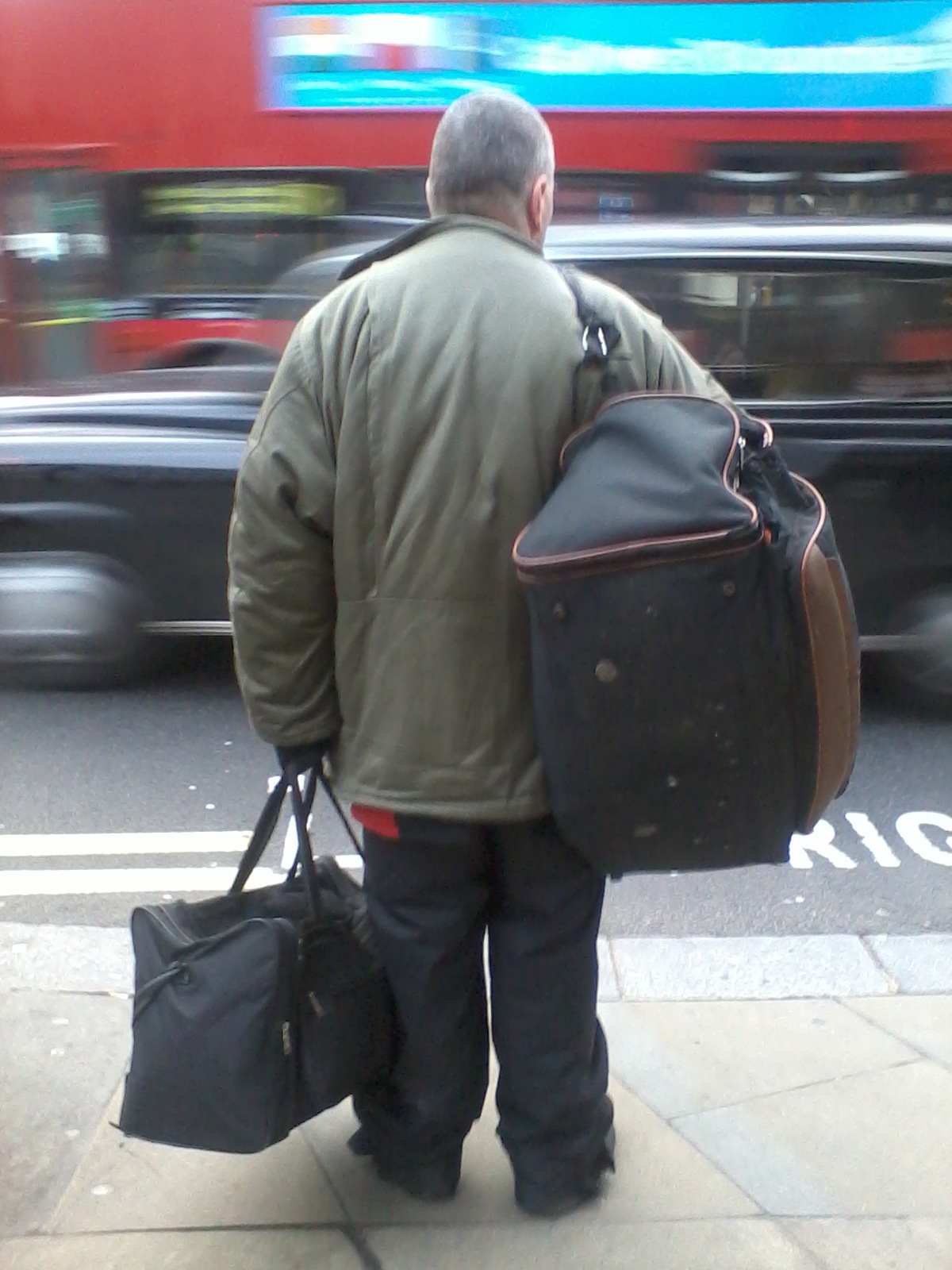 Back of a person standing on a city sidewalk, carrying two large duffel bags. The person is wearing a bulky olive-green jacket and dark pants. The background is blurred, showing the motion of city traffic, including a red double-decker bus. The overall impression is one of a traveler or someone possibly carrying belongings, possibly homeless, in a busy urban setting.