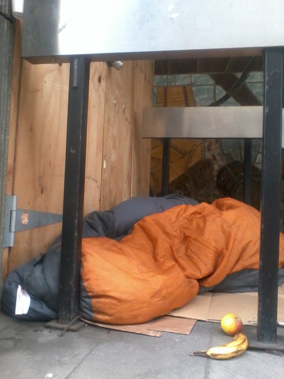 Person sleeping in a sleeping bag tucked under a metal structure, possibly outside.  A banana and an apple lie on the ground nearby. The overall impression is one of homelessness or poverty. The scene is bleak and suggests a lack of shelter and resources.