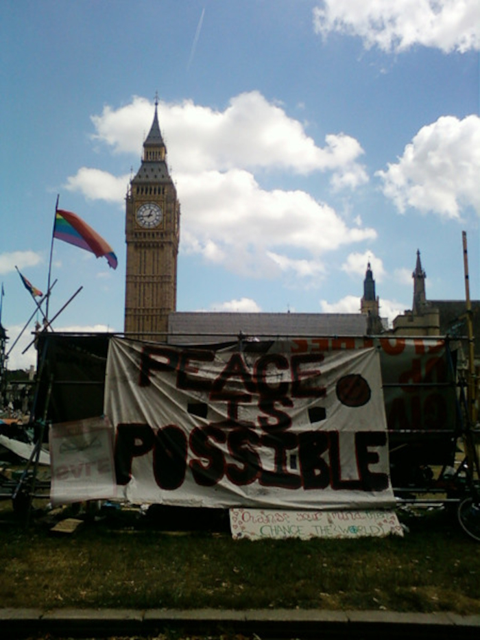 Protest scene in front of the Houses of Parliament in London. A large banner proclaims PEACE IS POSSIBLE in bold lettering. A rainbow flag is visible, suggesting a possible connection to LGBTQ+ rights or broader social justice themes. The overall context suggests a peaceful demonstration advocating for peace and possibly other related causes. The backdrop of the iconic Big Ben adds a significant visual element to the scene.