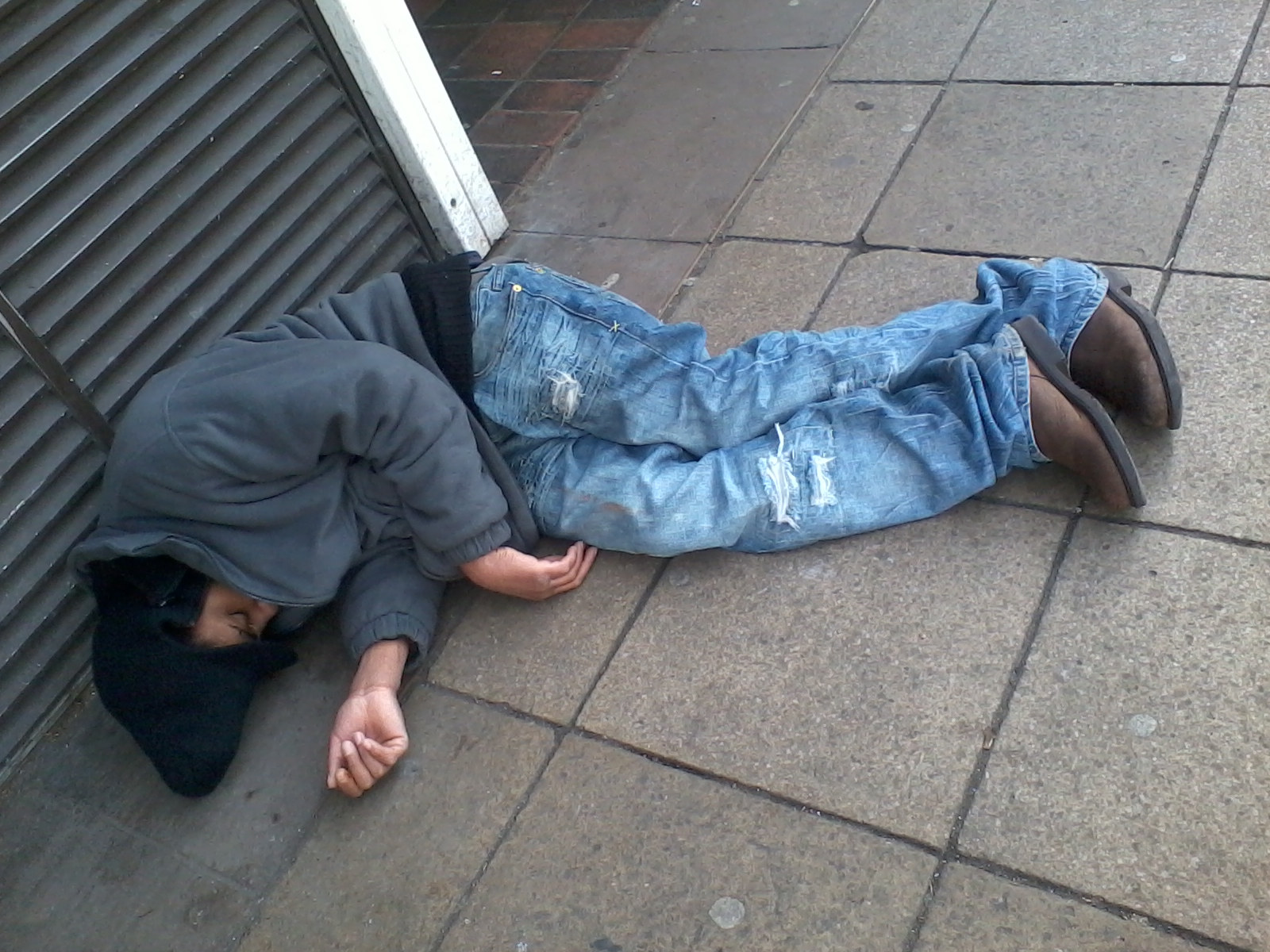 Person lying asleep or unconscious on a sidewalk. They are wearing a grey hooded jacket and blue jeans with rips and are wearing brown boots. The individual appears to be homeless or in a vulnerable state. The image is stark and lacks a narrative beyond depicting this scene of apparent destitution. The setting is an urban street.