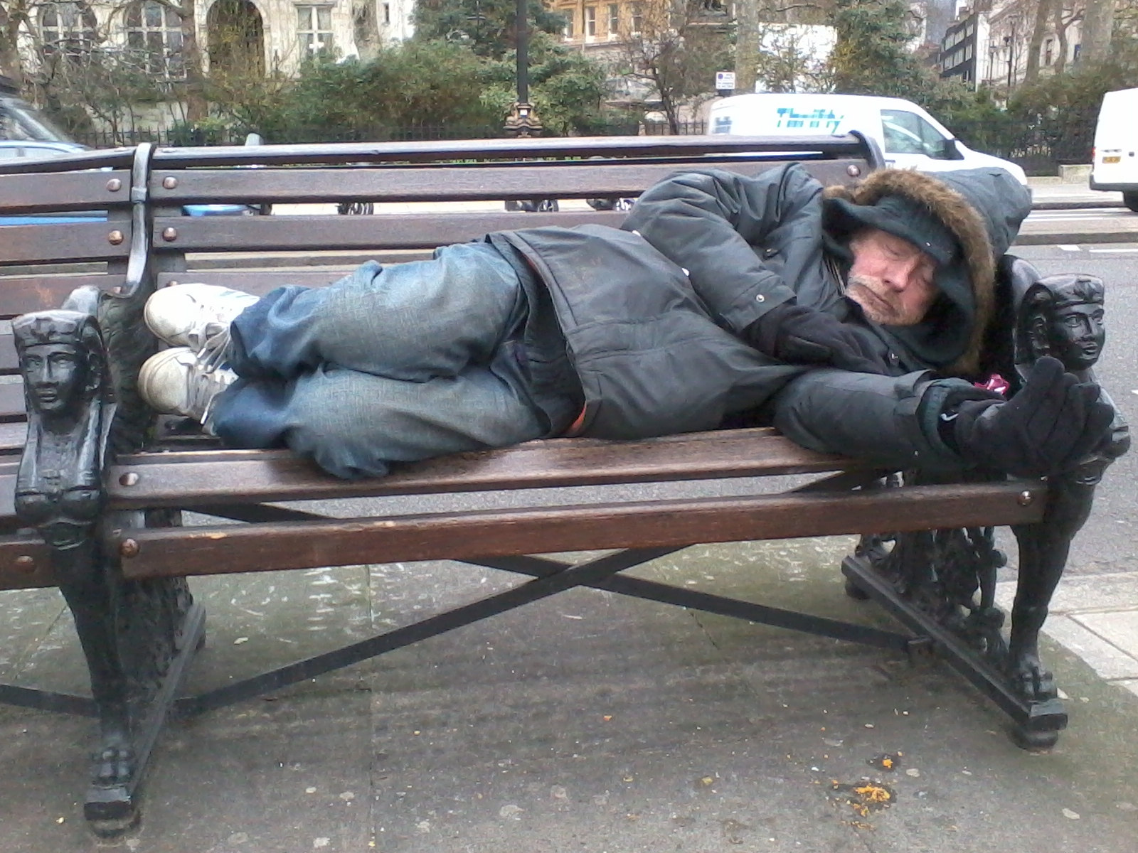 Homeless man sleeping on a park bench. He is wearing a dark-coloured jacket and jeans. He appears to be bundled up against the cold. The bench is made of wood and metal, and is ornately decorated at the ends. The background shows a city street with buildings and vehicles. The overall impression is one of poverty and hardship.