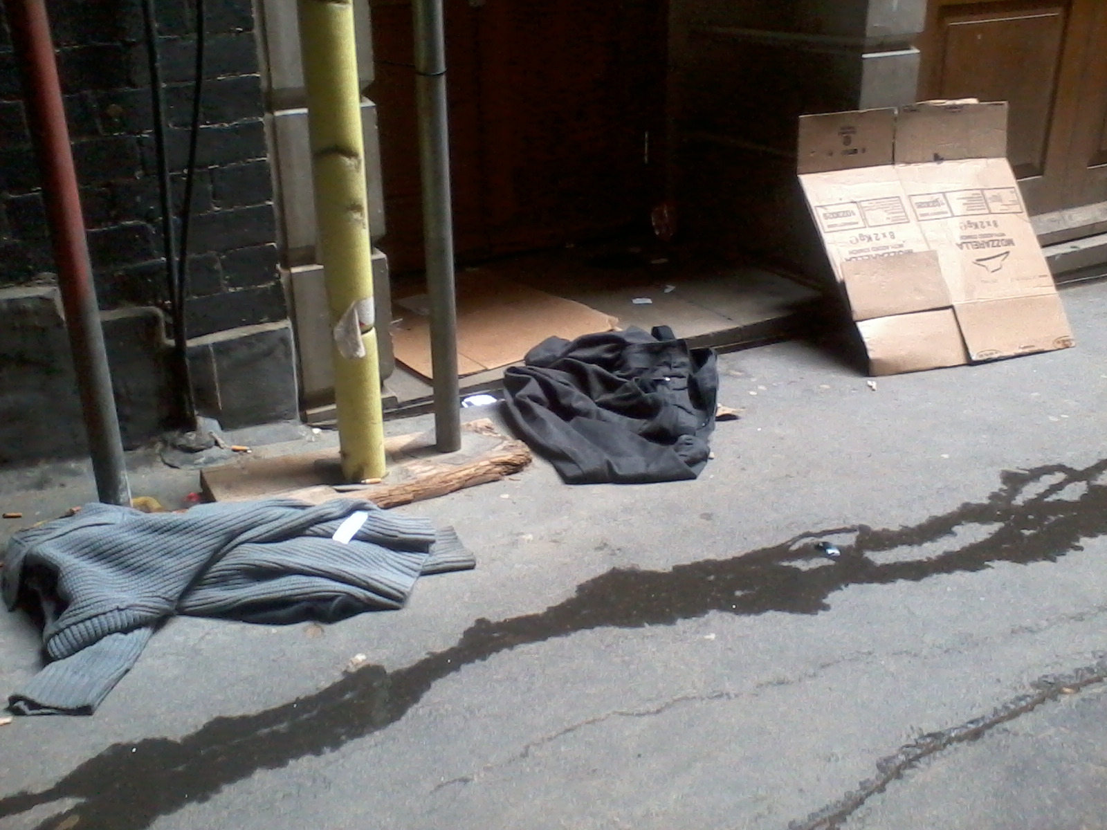 Dark alleyway with discarded clothing and cardboard boxes on the ground. There are two piles of clothing, one a dark-colored pair of trousers and the other a grey sweater. A piece of cardboard leaning against the wall appears to be part of a larger box. The overall impression is one of poverty and homelessness.