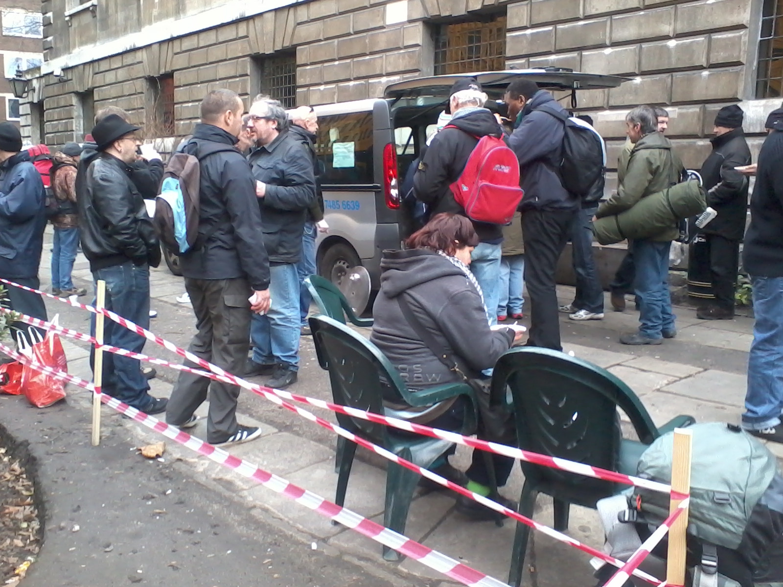 Group of people, mostly men, standing and sitting outside a building. They appear to be waiting, possibly for transportation, as a van with its rear door open is visible. Many are carrying backpacks and bags, suggesting a journey or displacement. The setting seems urban, with a stone building forming the backdrop. A red and white caution tape is present, possibly indicating a designated waiting area or restricting movement. The overall mood is somewhat somber and suggestive of a temporary situation. The picture might depict refugees or migrants waiting for assistance or resettlement.