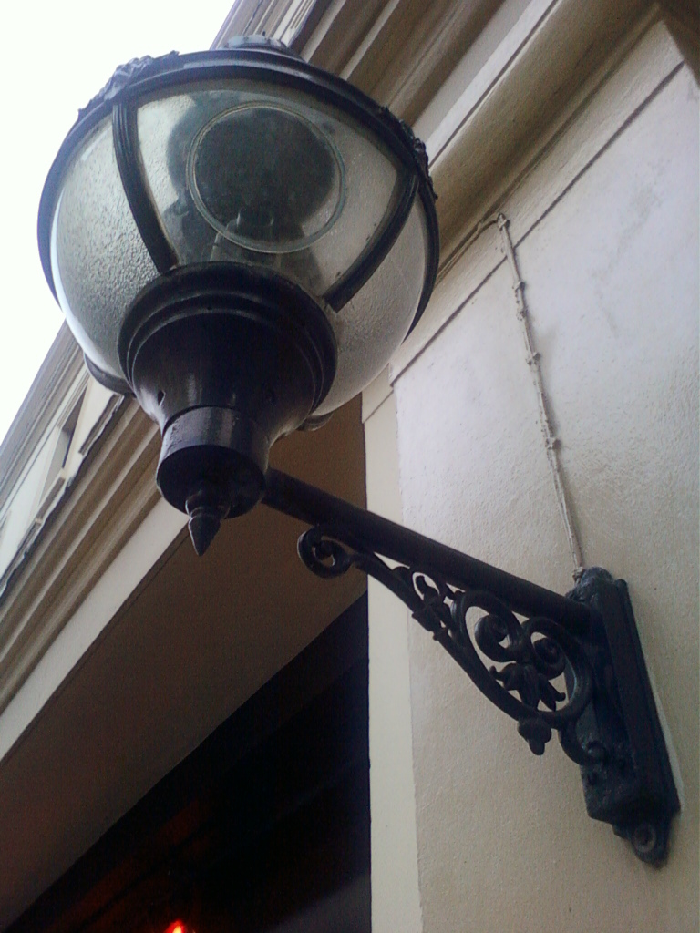 Close-up view of an ornate, dark-coloured wall-mounted lamp. The lamp has a rounded, glass globe with a textured surface and a decorative metal bracket featuring swirling, floral designs. The lamp is affixed to a light-colored building's exterior wall. A hint of a dark interior space is visible behind the lamp.