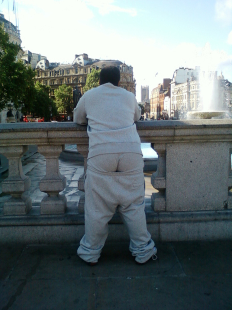 Person wearing grey sweatpants, leaning on a stone balustrade overlooking a city square with a fountain. The background features several buildings and some trees. The overall impression is one of quiet observation or contemplation. The person's clothing is somewhat baggy and the focus is on their posture and the urban scene behind them.