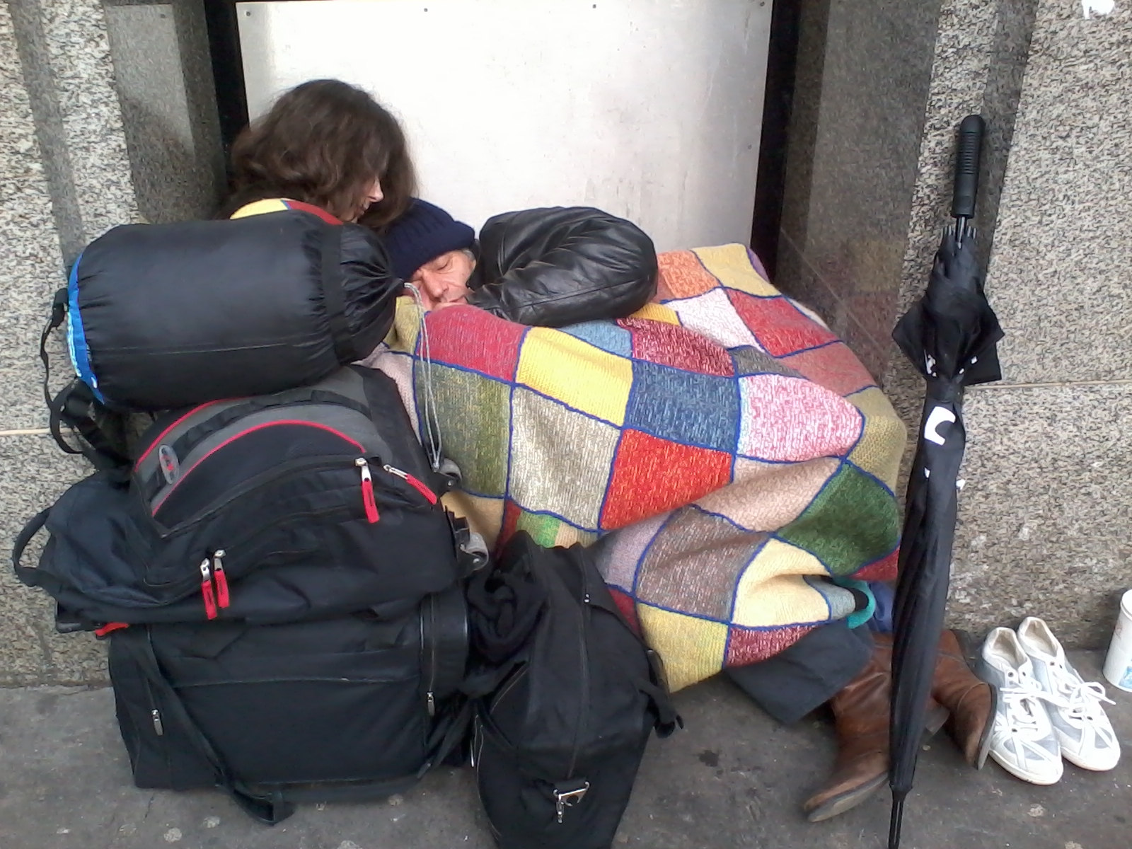 Homeless couple huddled together for warmth in a doorway. A young woman with long brown hair rests her head on the shoulder of an older man, who is wrapped in a patchwork blanket. Surrounding them are several backpacks and bags, suggesting all their belongings. A closed umbrella leans against the wall nearby. The scene evokes a sense of vulnerability and hardship, highlighting the realities of homelessness and the need for compassion.