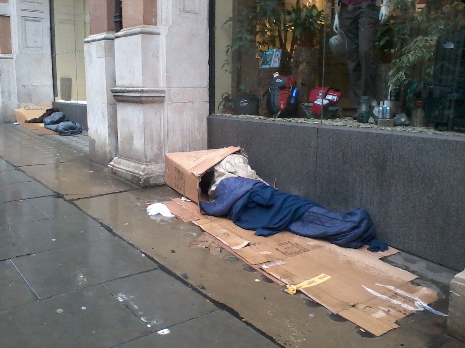 Homeless person sleeping on the street, in a cardboard box. The person is wrapped in a blue blanket. The setting appears to be a city street near a shop window. Another person's belongings are visible a short distance away, suggesting others may also be experiencing homelessness in the area. The overall impression is one of poverty and hardship.