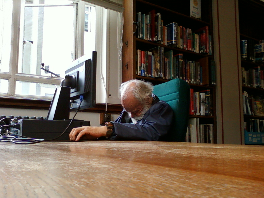 Elderly man with long white hair and a beard, slumped over a desk in front of a computer monitor. He appears to be asleep or resting his head on the desk. He's wearing a dark jacket and a watch. The setting appears to be a library or study, with bookshelves filled with books visible in the background. The overall mood is one of quiet rest or perhaps weariness.