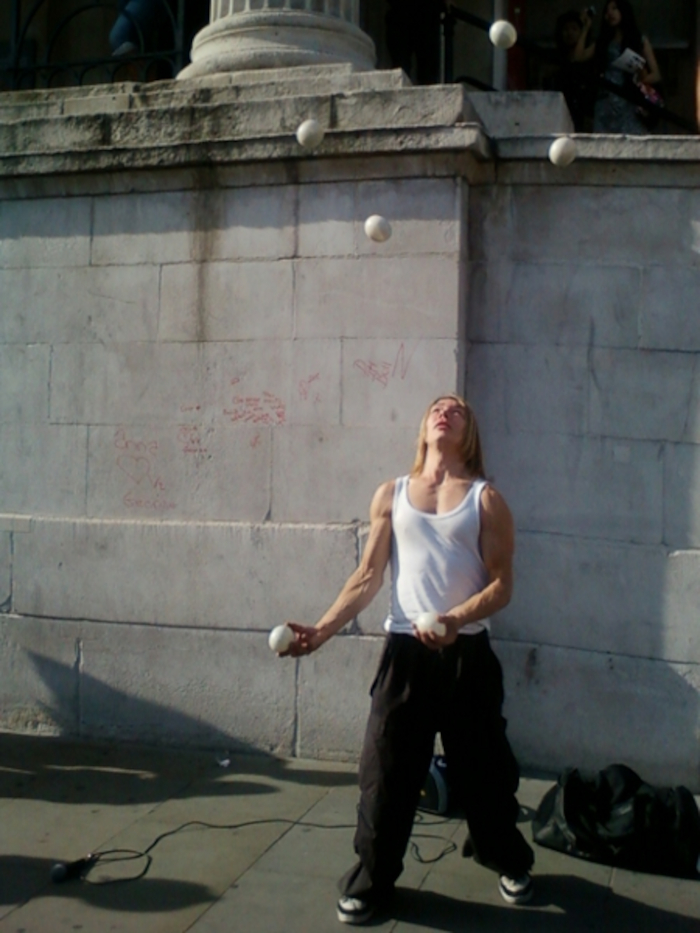 Person juggling five white balls in front of a stone wall. The juggler is a person with shoulder-length blonde hair, wearing a white tank top and dark pants. The background includes a stone wall with graffiti and some people in the distance.