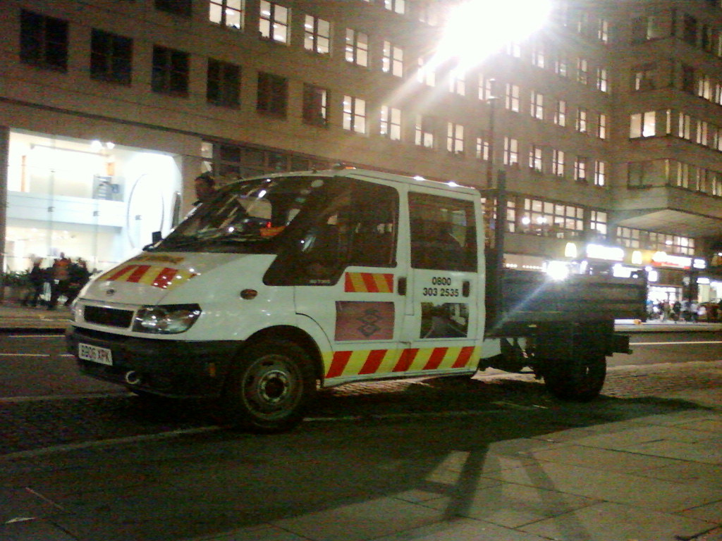 White flatbed truck parked on a city street at night. The truck has red and yellow diagonal stripes on its sides and a number plate. The background features a multi-story building with illuminated windows and a fast-food restaurant. The scene is dimly lit, suggesting it was taken at night. The overall impression is of an ordinary street scene in a city, possibly showing a utility or service vehicle.