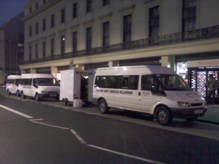 Three white vans parked at night along the side of a large, classic-looking building. One van is smaller, possibly a cargo van, and appears to be hitched to a larger passenger van. The building seems to be in a city center, possibly in the UK, given the architectural style. The overall impression is of a nighttime delivery or service related activity.