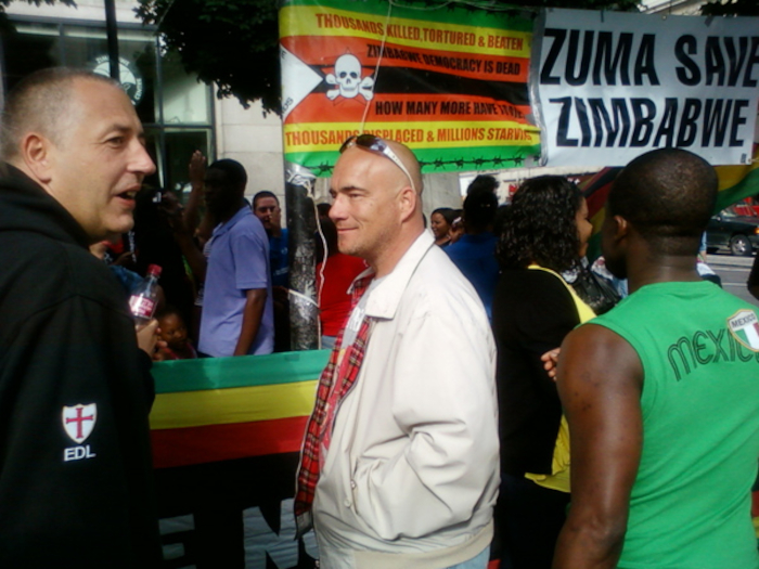 Protest or demonstration, likely related to the political situation in Zimbabwe. A prominent sign reads ZUMA SAVE ZIMBABWE, indicating a plea for intervention from South African President Jacob Zuma. Another sign details the suffering within Zimbabwe, citing killings, torture, displacement, and starvation. The photo captures a moment of interaction between two men: one wearing a black jacket with EDL (likely the English Defence League) insignia, and the other, a lighter-complexioned man in a cream jacket. The contrast in their attire and likely affiliations suggests a potential clash of ideologies or perspectives within the event. The background features a diverse group of protesters, some holding Zimbabwean flags. 