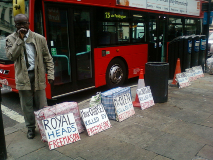 Dark-skinned man standing beside a red double-decker bus in what appears to be a London street. He is dressed in a beige jacket and light-coloured trousers, and he appears to be covering his mouth with his hand. At his feet are several bags and numerous hand-made signs. These signs allege that several individuals were killed by Freemasons, naming specific individuals. The scene suggests a protest or demonstration related to conspiracy theories surrounding the Freemasons. The overall impression is one of a solitary individual expressing a strong, possibly controversial, belief. The image's mood is somber and somewhat unsettling.