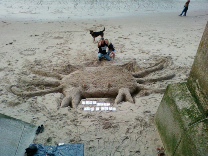 Large sand sculpture of a crab on a beach. Two people are sitting in front of it, and a dog is standing nearby. There are also some small pieces of paper scattered in front of the sculpture, which seem to be handwritten notes. In the background, a person is walking away on the beach. The overall scene suggests a fun, playful activity on a beach, possibly a collaborative art project.