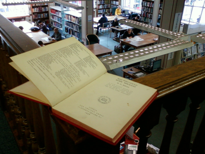 Open book resting on a railing overlooking a library. The book appears to be a catalogue or index, with densely packed text visible on the open pages. The library itself is bustling with activity, showing numerous people seated at tables, presumably reading or studying. The overall setting suggests a quiet but active space of learning and research. The contrast between the intimate view of the book and the broader view of the library creates a sense of both individual focus and communal learning.