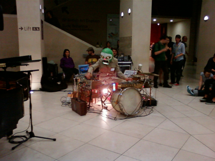 Person wearing a mask that resembles a skull performing a musical act in a large, open space. They are surrounded by various musical instruments and electronic equipment. A small audience is visible in the background. The setting appears to be a modern, public building, perhaps a gallery or museum.