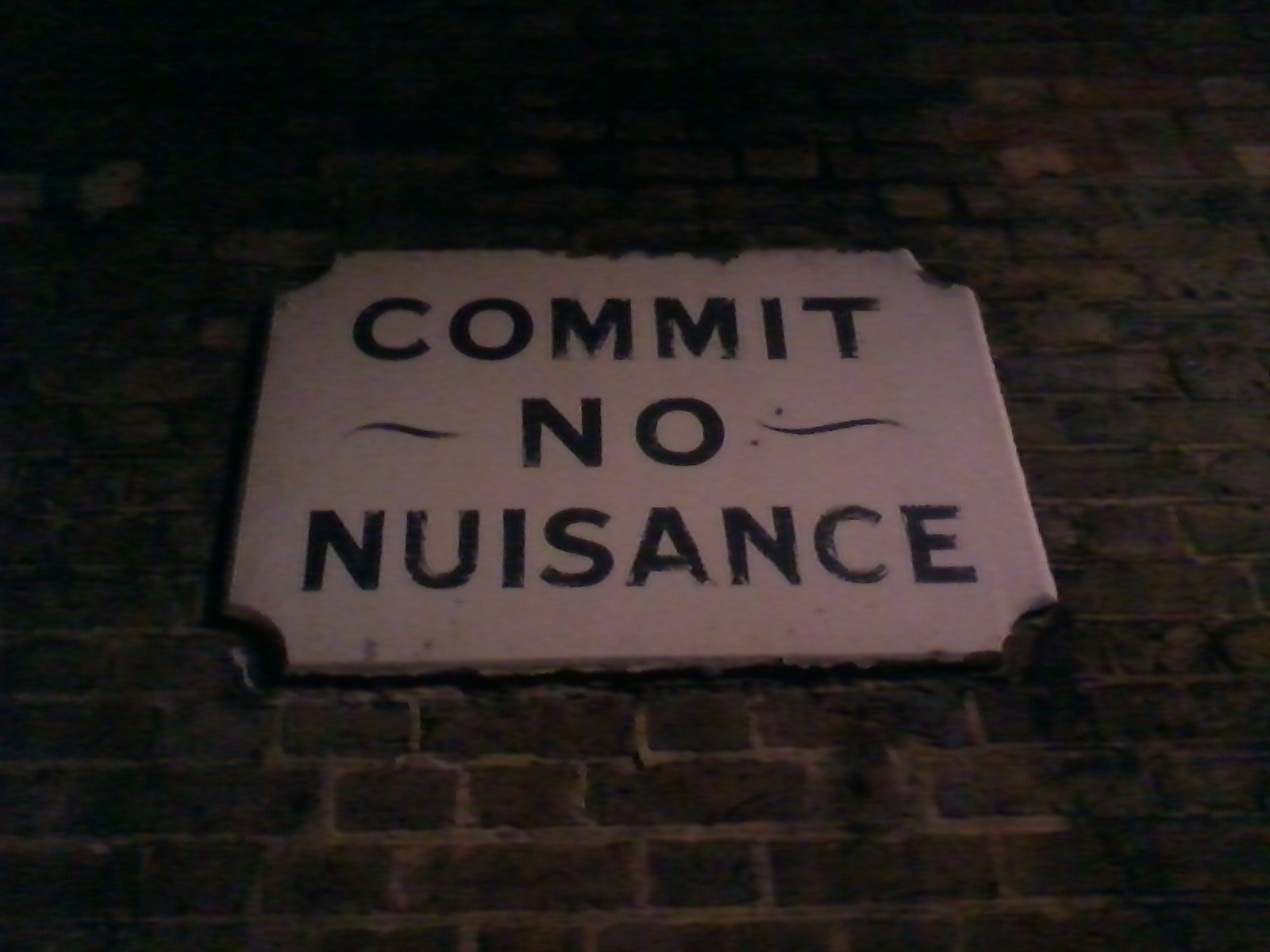 Weathered, rectangular sign affixed to a brick wall. The sign, which appears to be made of stone or plaster, displays the words COMMIT NO NUISANCE in bold, uppercase, black lettering against an off-white background. The NO is slightly smaller than the other words and has a small decorative flourish underneath it. The overall impression is that of an old, possibly historical, sign conveying a clear and concise instruction or warning.