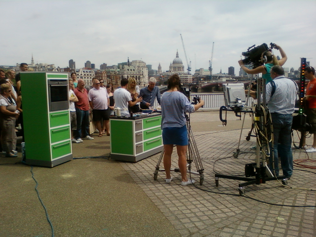 Television cooking show being filmed outdoors, likely in London, given the backdrop of St Paul's Cathedral and the River Thames. A small audience stands behind two bright green kitchen units, while a camera crew films a chef preparing food. The scene is bustling with activity, suggesting a live or on-location broadcast.