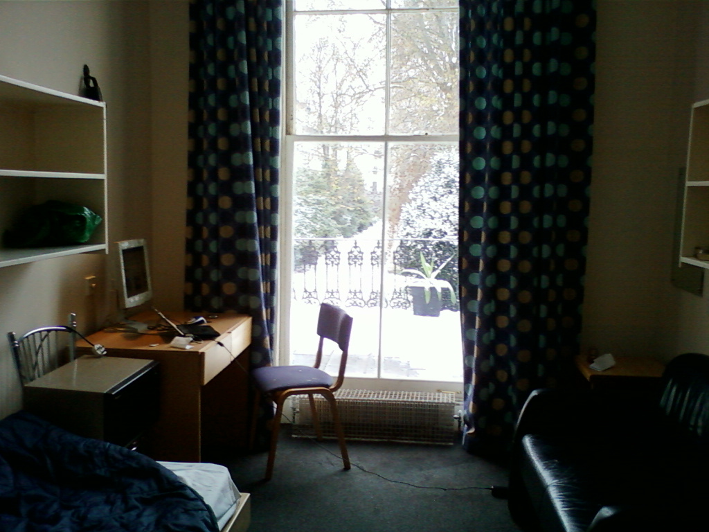 Hostel room, viewed from the corner. A desk with a computer is positioned in front of a large window draped with patterned curtains. Snow is visible outside. A chair is tucked under the desk, and a bed partially occupies the foreground. There are shelves on the walls holding personal items. The room has a somewhat minimalist feel. The overall mood is quiet and contemplative.