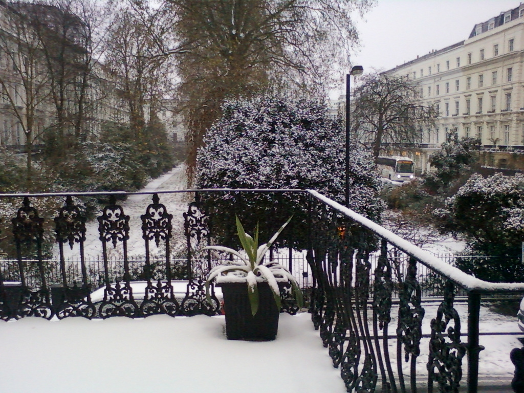 Snow-covered scene viewed from a balcony. A wrought iron balcony railing is in the foreground, with a potted plant sitting on the snow-covered balcony. Beyond the railing, a snow-dusted garden path leads between rows of snow-covered trees and buildings. A bus is partially visible in the distance. The overall impression is one of a quiet, wintry day in an urban environment.