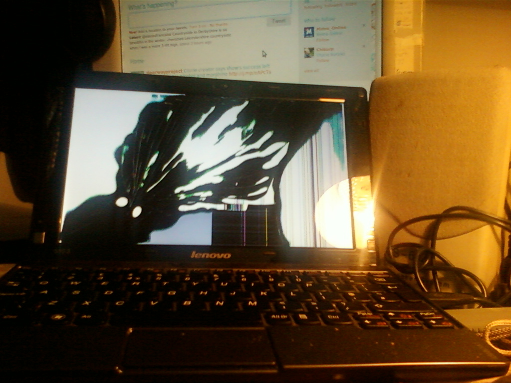 Lenovo laptop with a severely cracked screen. The crack is extensive and displays a pattern of vertical lines and distortions across a majority of the screen. The laptop is illuminated from the right side. Part of a computer monitor and speaker are visible in the background. The overall impression is one of damage and malfunction.