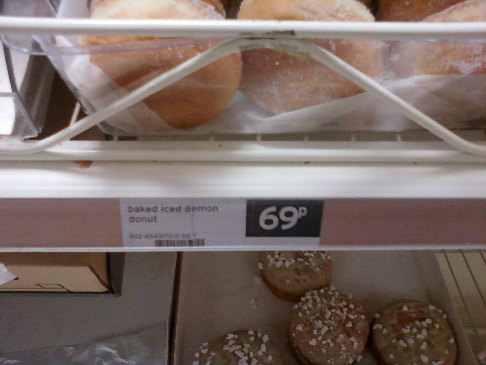 Close-up of a shelf in a store displaying baked iced demon doughnuts. The doughnuts are in a clear plastic container. A price tag indicates they cost 69 pence. Below the shelf, more doughnuts, possibly the same kind but with white sprinkles, are visible. The overall lighting is dim, and the image quality is somewhat blurry.