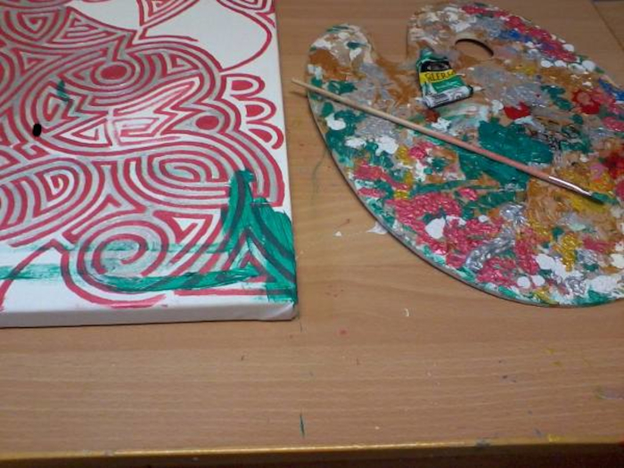 Canvas with an abstract design. The design consists primarily of swirling, concentric lines in red and silver, with a small area of green paint added in the bottom right corner. Beside the canvas is a paint palette with a variety of colors, including red, green, gold and silver, and a paintbrush resting on it. The overall impression is one of a work in progress.