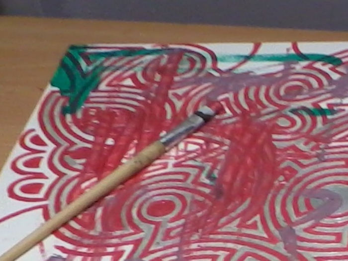 Artwork in progress. A paintbrush rests on a piece of paper or canvas covered in a predominantly red and silver swirling, concentric circular pattern. There are also streaks of green paint visible. The overall impression is one of energetic, possibly abstract, expression. The paint appears to be applied somewhat haphazardly, suggesting a spontaneous creative process.