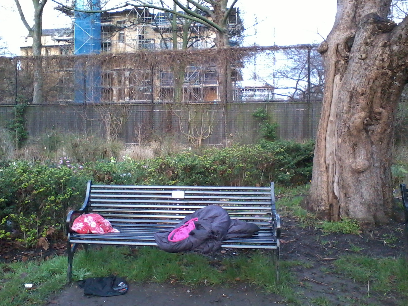 Park bench in a somewhat neglected state. On the bench are discarded items: a pink and red patterned fabric, and a dark grey sleeping bag partially obscuring a pink item underneath. A dark object lies on the ground near the bench's base. The background features a chain-link fence separating the park from a building under construction, which is partially obscured by overgrown vines. The overall impression is one of urban decay and possible homelessness.