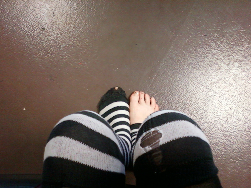 Leonie's legs from the knees down, wearing black and grey striped knee-high socks. One sock has a noticeable hole in it. The feet are bare except for the socks, and rest on a dark brown textured floor. The overall impression is casual and slightly informal.