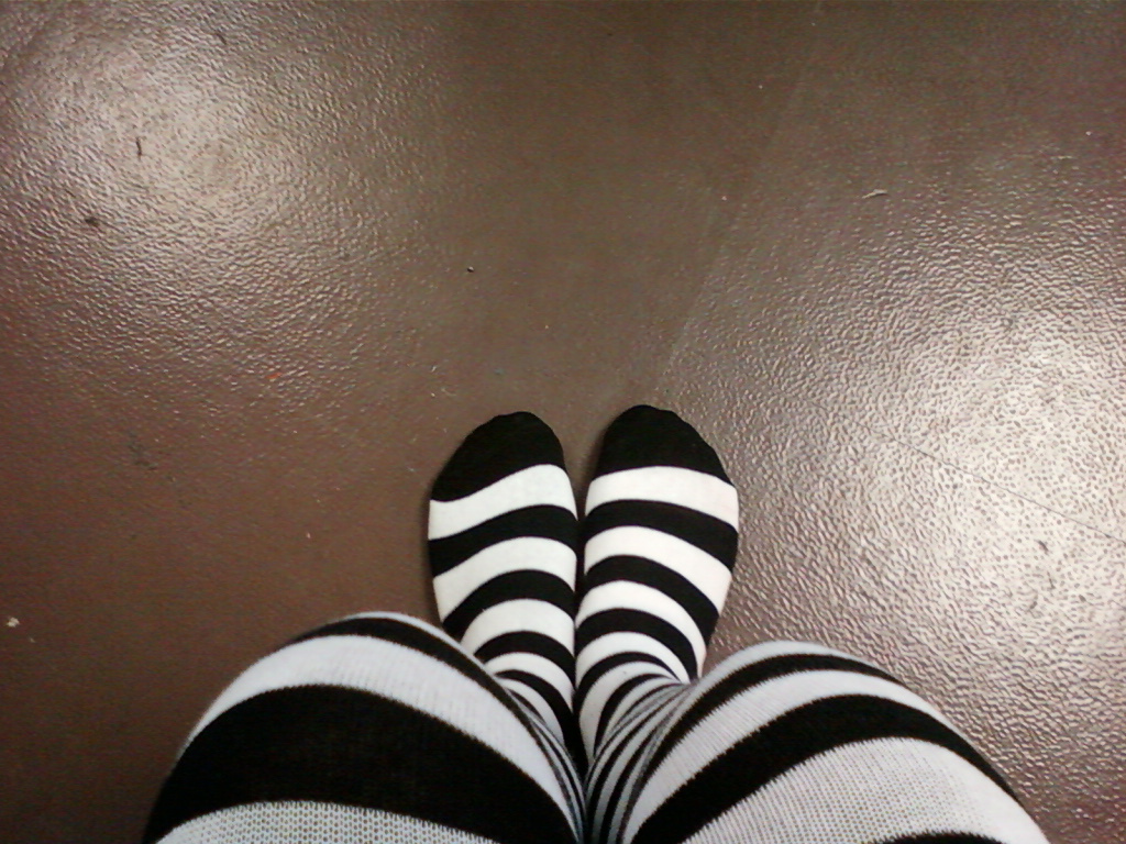 Close-up, from a slightly elevated angle, of a person's legs clad in black and white striped knee-high socks. The socks are prominent, and the background is a textured brown floor. The focus is sharply on the socks and legs. The overall impression is simple and somewhat minimalist.