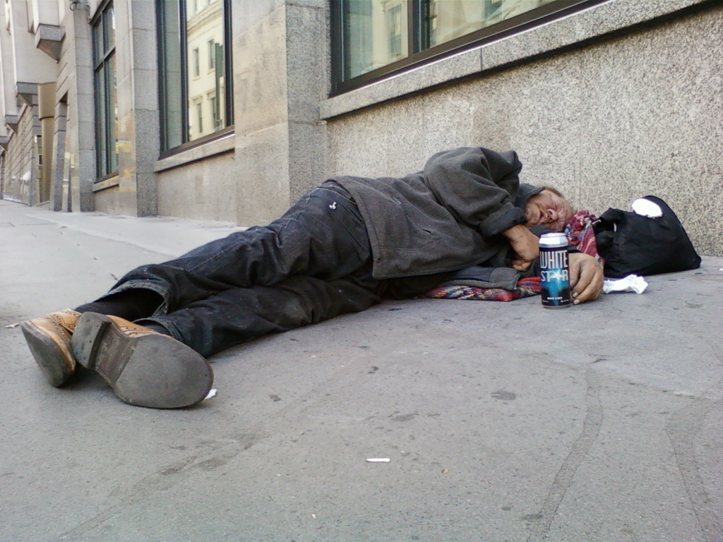 Homeless man asleep on the sidewalk next to a building. He is lying on his side, his body partially covered by a blanket.  His belongings, including a bag, are next to him. A can of White Star beer is visible near his hand. The overall look is one of poverty and hardship. The image has a stark, realistic quality.