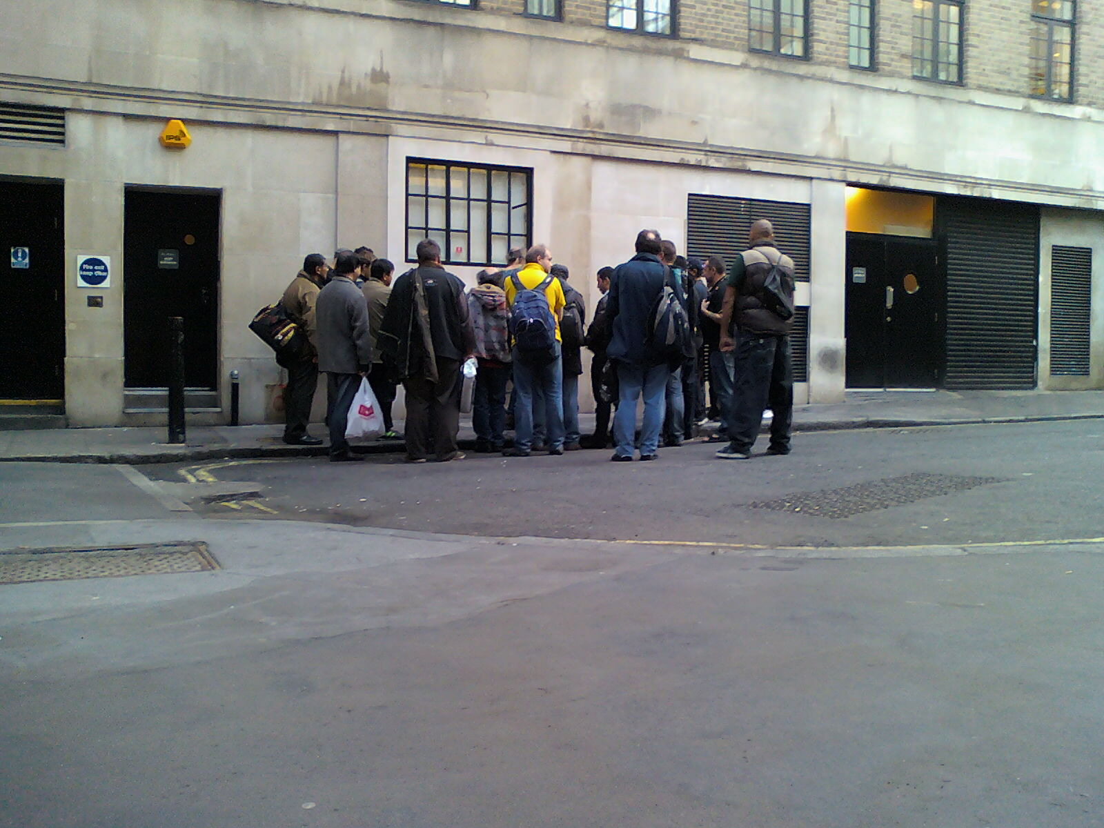 Group of approximately twenty people standing outside a building. They appear to be waiting for something or someone. The setting appears to be a city street in what looks like London, UK, based on the architecture of the building. The individuals are dressed in casual clothing, and there is little to distinguish them individually. The overall mood is one of quiet expectation or perhaps mild anticipation. The image lacks strong narrative or emotional elements beyond the implied sense of waiting.