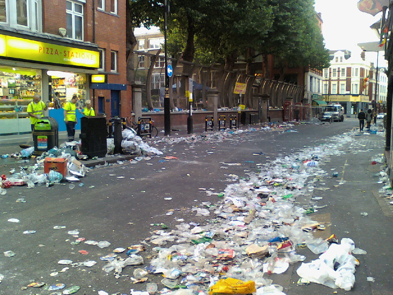Street scene, heavily littered with trash. Empty plastic bottles, cans, and other refuse are strewn across the roadway and sidewalk. Several sanitation workers are visible, apparently beginning the process of cleaning up the mess. The background shows buildings and a relatively quiet street. The overall impression is one of post-event cleanup, perhaps after a celebration or festival.