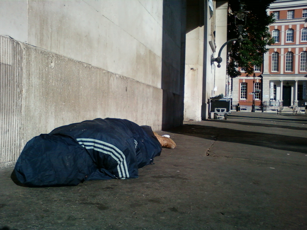 Person lying face down on the ground, curled up in a dark blue jacket with light grey stripes on the sleeves, near a wall. Their feet are visible, and one shoe is partially seen. The setting appears to be an urban area, with a building and street in the background. The overall impression is one of homelessness or vulnerability.