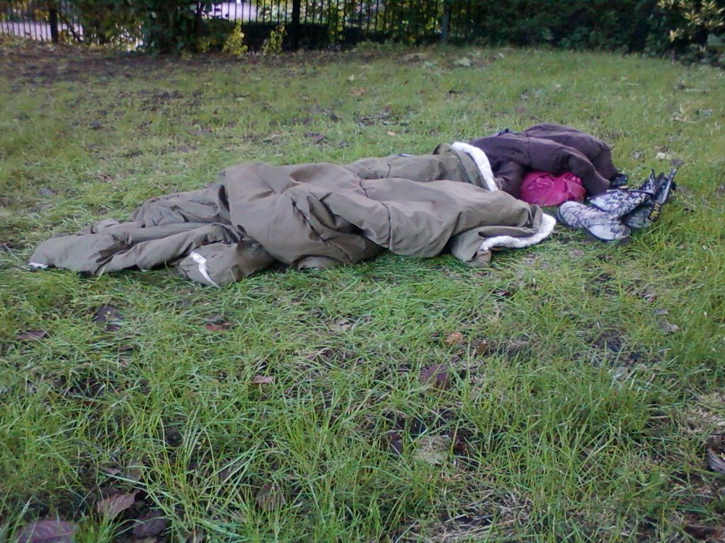 Person sleeping outdoors on the grass. They are wrapped in a sleeping bag and other clothes, including a jacket and what appears to be a pair of boots. The setting looks like a park or other green space. The overall impression is one of homelessness or rough sleeping.