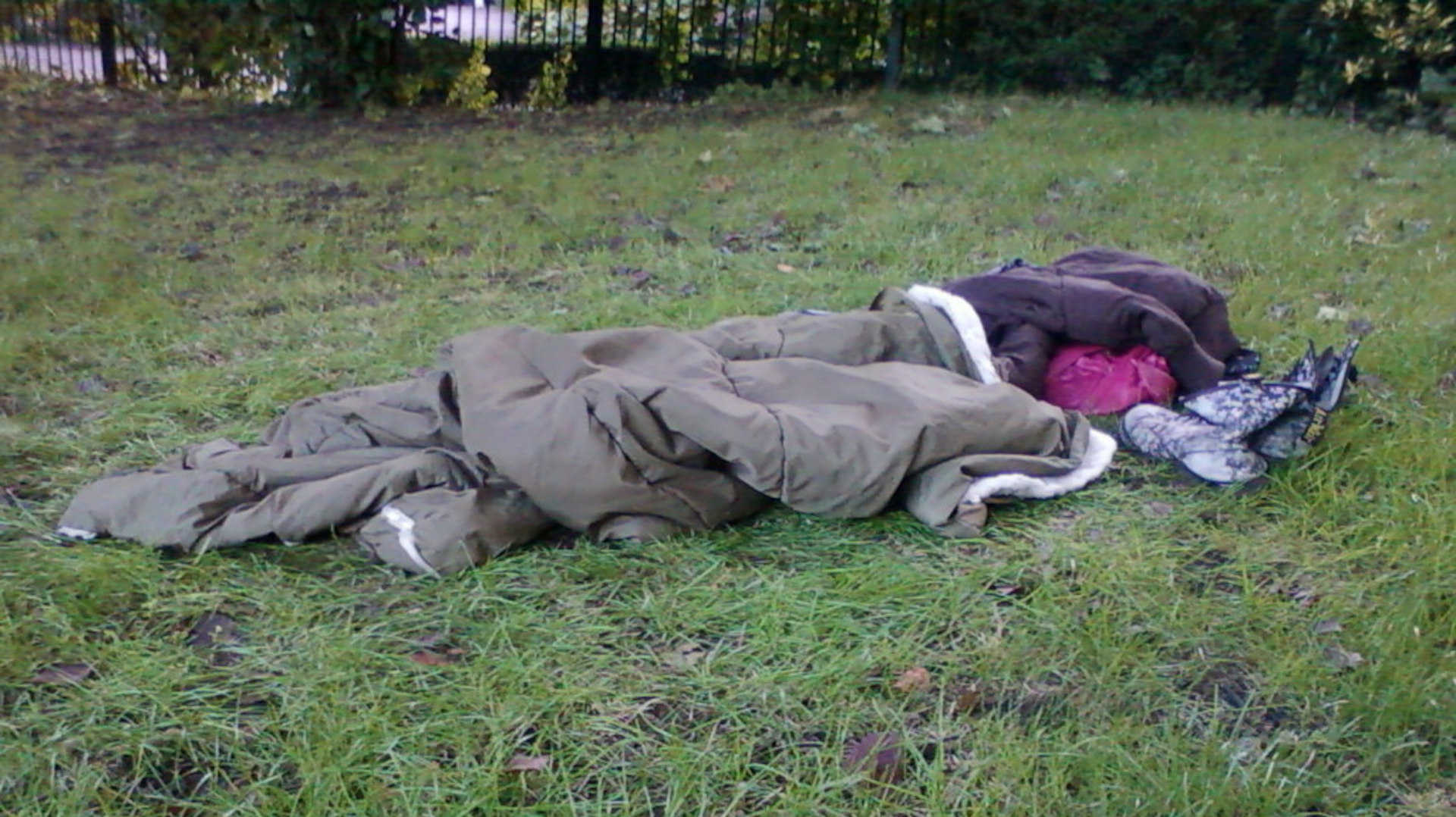 Leonie sleeping outdoors, wrapped in a large olive-green sleeping bag. Near the head of the sleeping bag are additional clothes and footwear. The setting appears to be a grassy area, possibly a park or common land. The overall impression is one of homelessness or rough sleeping.