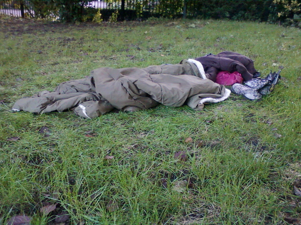 Leonie sleeping outdoors, wrapped in a large olive-green sleeping bag. Near the head of the sleeping bag are additional clothes and footwear. The setting appears to be a grassy area, possibly a park or common land. The overall impression is one of homelessness or rough sleeping.