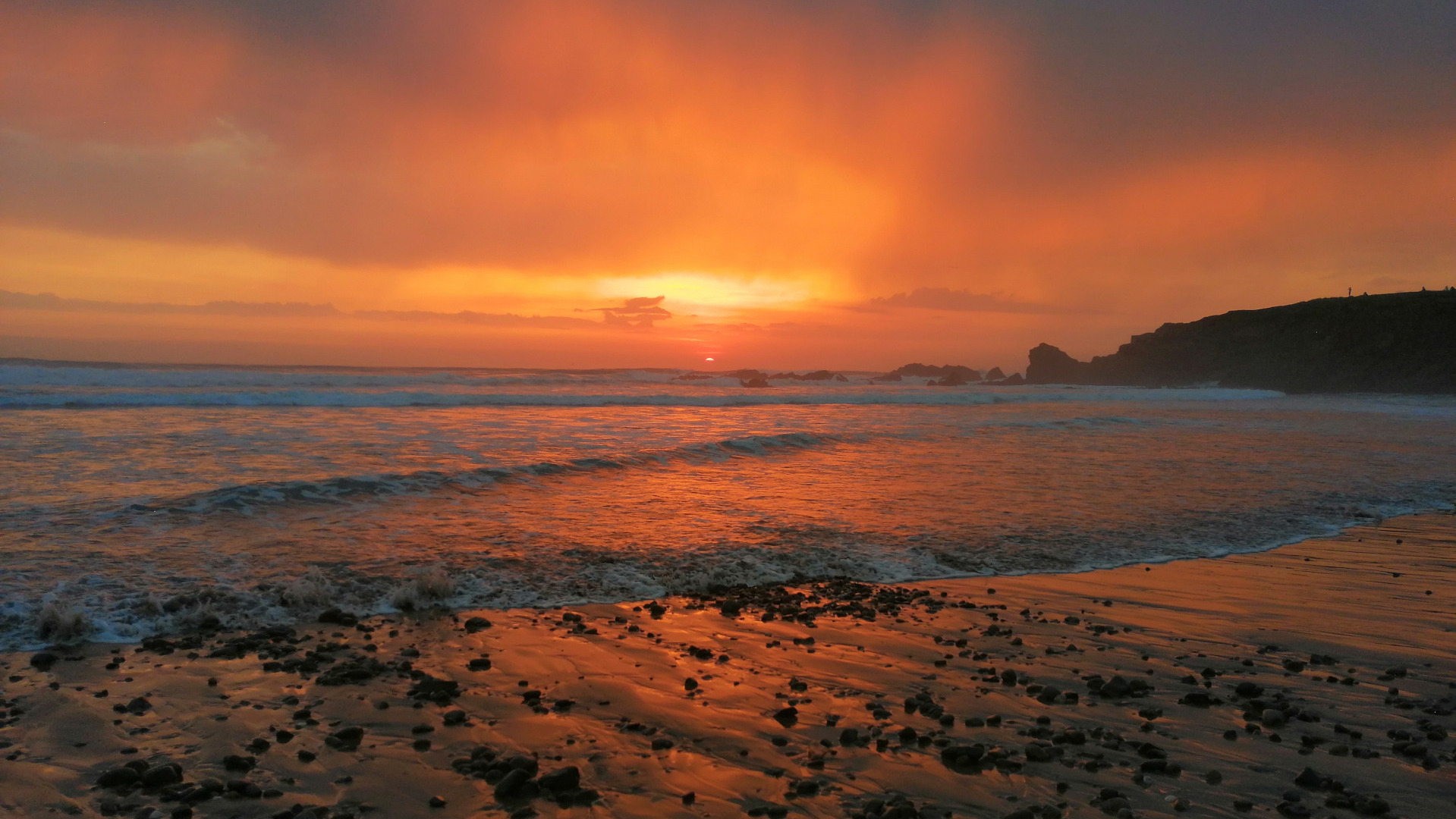 A beach scene at sunset. The sky is a vibrant orange and red, reflecting in the wet sand and the waves of the ocean. Dark rocks are scattered across the beach in the foreground. In the distance, a dark shoreline is silhouetted against the bright sky. The overall impression is one of dramatic, warm beauty.