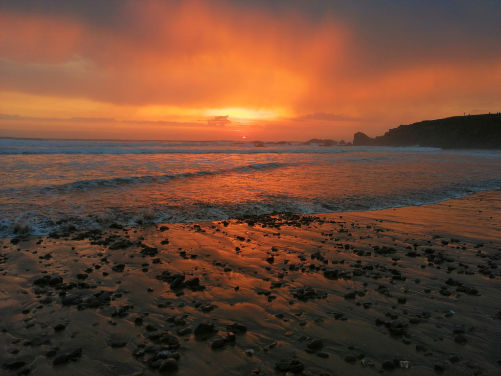 Beach scene at sunset. The sky is a vibrant orange and red, reflecting in the wet sand and the waves of the ocean. Dark rocks are scattered across the beach in the foreground. In the distance, a dark shoreline is silhouetted against the bright sky. The overall impression is one of dramatic, warm beauty.