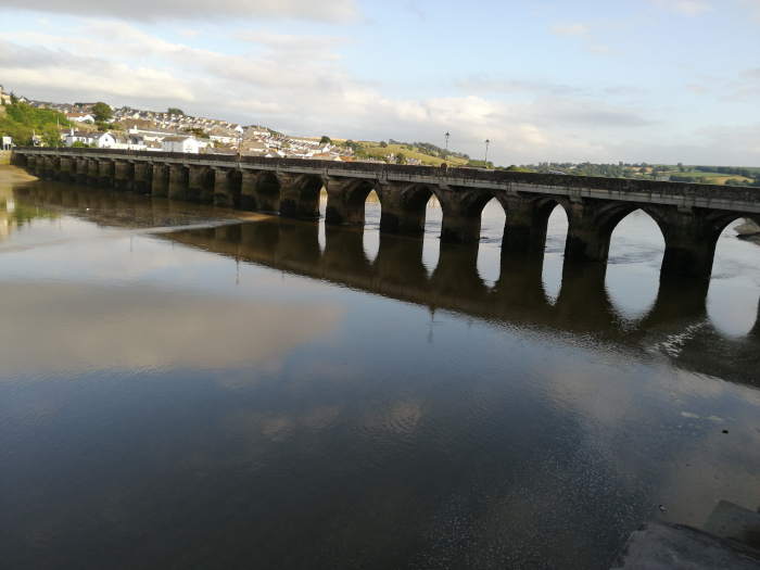 Long stone bridge spanning a calm river. The bridge has numerous arches and appears old. Beyond the bridge, a small town or village is visible on a hillside. The sky is mostly cloudy, and the river reflects the bridge and the sky. The overall impression is one of peaceful tranquillity.
