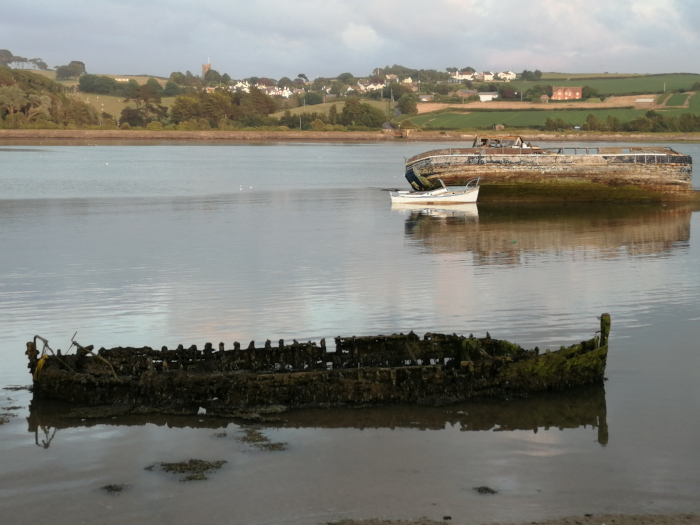 Calm body of water with a tranquil landscape in the background. In the foreground are two wrecked boats: one largely intact but decaying, covered in algae and partially submerged, and a smaller, more decomposed wreck. A small, seemingly functional boat is moored near the larger wreck. The background features a picturesque village nestled amongst rolling hills. 